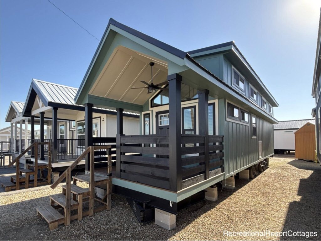Modern tiny house with teal siding, black trim, and a covered porch. Features large windows, pitched roof, and ceiling fan under a clear blue sky.