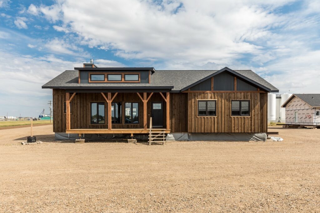 A rustic wooden house with large windows and a small porch stands on a gravel lot under a blue sky with scattered clouds, conveying a cozy rural feel.
