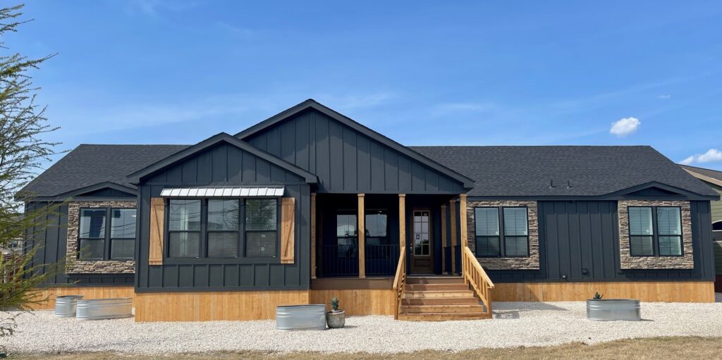 Single-story, modern manufactured home with a dark exterior and stone accents. Wooden steps lead to the central porch. Clear blue sky above.
