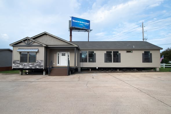A beige modular home with a stone-trimmed porch and a prominent white door sits under a partly cloudy sky. A billboard is visible above the house.