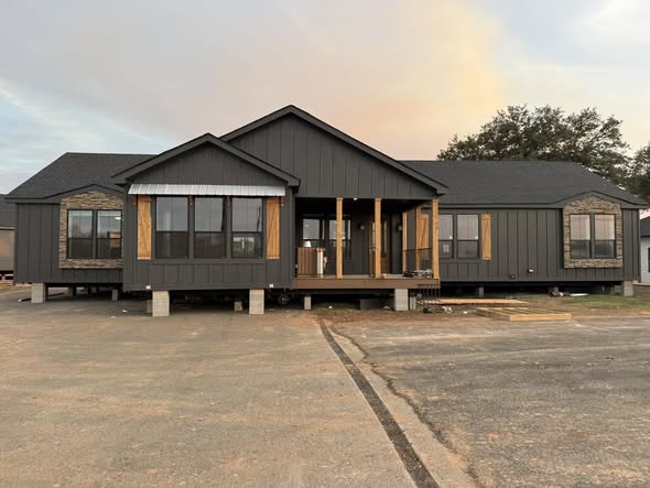 A modern, dark-colored house with large windows, stone accents, and a welcoming porch sits under a soft sky at sunset, conveying a serene atmosphere.
