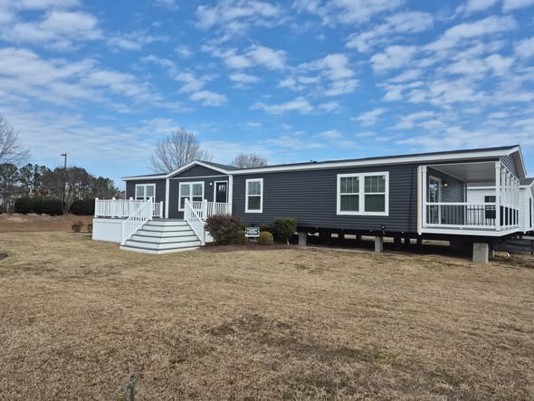 A modern, gray mobile home with white trim and a front porch sits elevated on a grassy lot under a partly cloudy blue sky, evoking a peaceful, rural atmosphere.