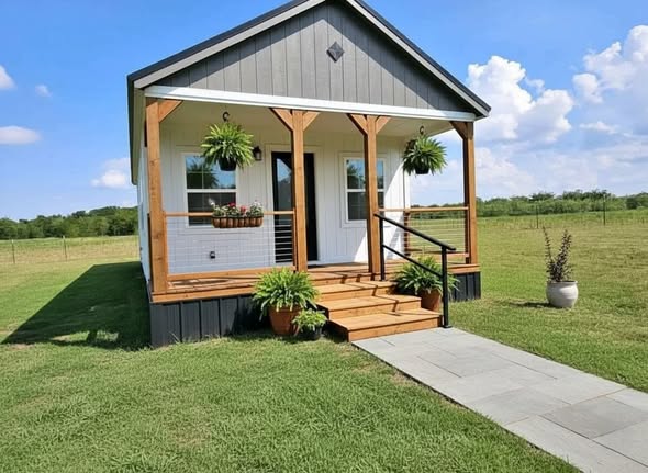 A small, charming gray house with a wooden porch sits in a lush green field under a blue sky. Hanging plants and potted ferns adorn the entrance.