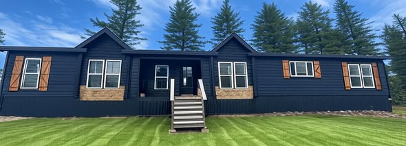 A modern, navy blue house with large windows and wooden shutters stands on a manicured lawn. Pine trees and a bright blue sky form the scenic backdrop.
