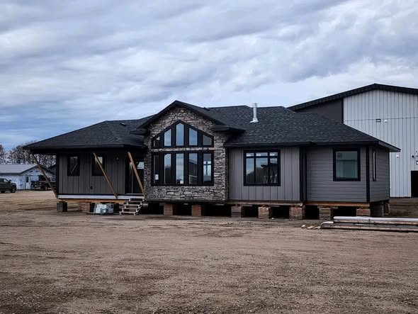 A modern gray house with stone accents is raised on blocks, indicating construction. Overcast sky and dirt ground create a work-in-progress atmosphere.