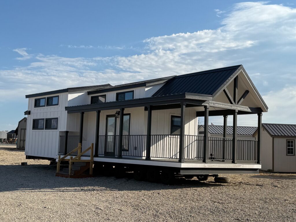 Modern tiny house with large windows and black trim under a blue sky. The house has a loft, a covered porch, and a metal roof, set on gravel.