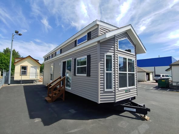 A compact, gray tiny house with a peaked roof and large windows sits in a sunny lot. Wooden steps lead to the entrance. The sky is clear with wispy clouds.