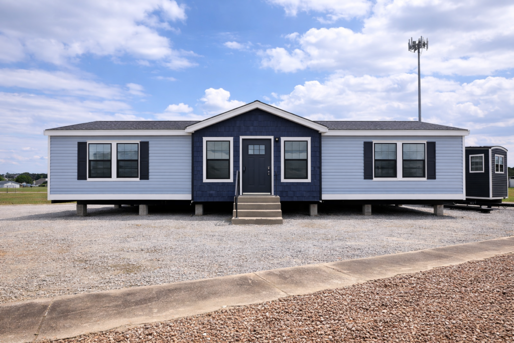 A blue and white manufactured home with a dark blue door and multiple windows sits on a gravel lot under a partly cloudy sky, conveying a sense of simplicity.