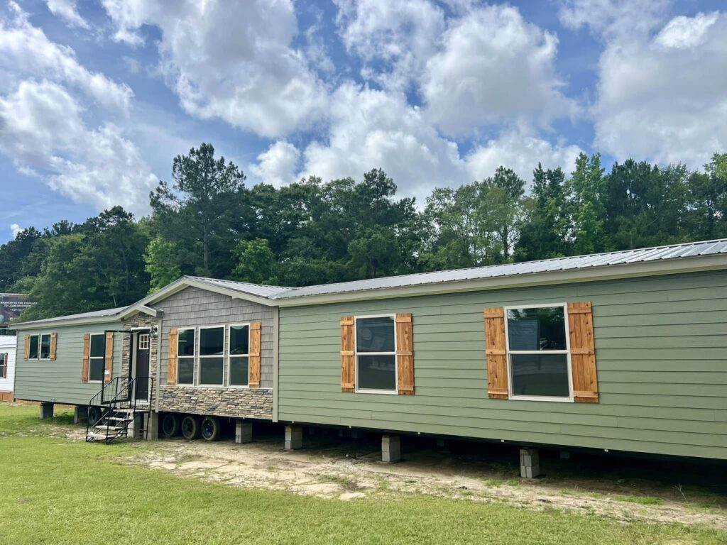 Green mobile home with wood shutters and stone accents, set on a grassy area with a backdrop of lush trees under a partly cloudy sky.