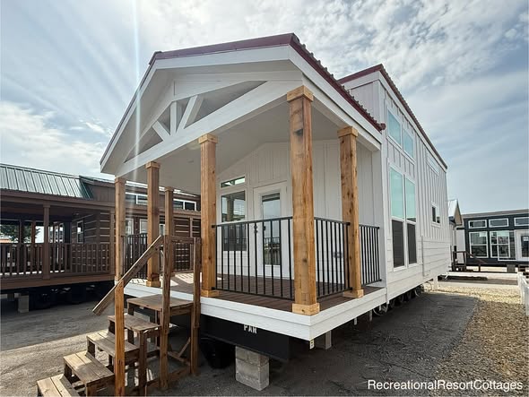 A modern tiny house with a gabled roof and wooden posts stands prominently. It has large windows, a small porch, and steps leading up. The sky is partly cloudy.