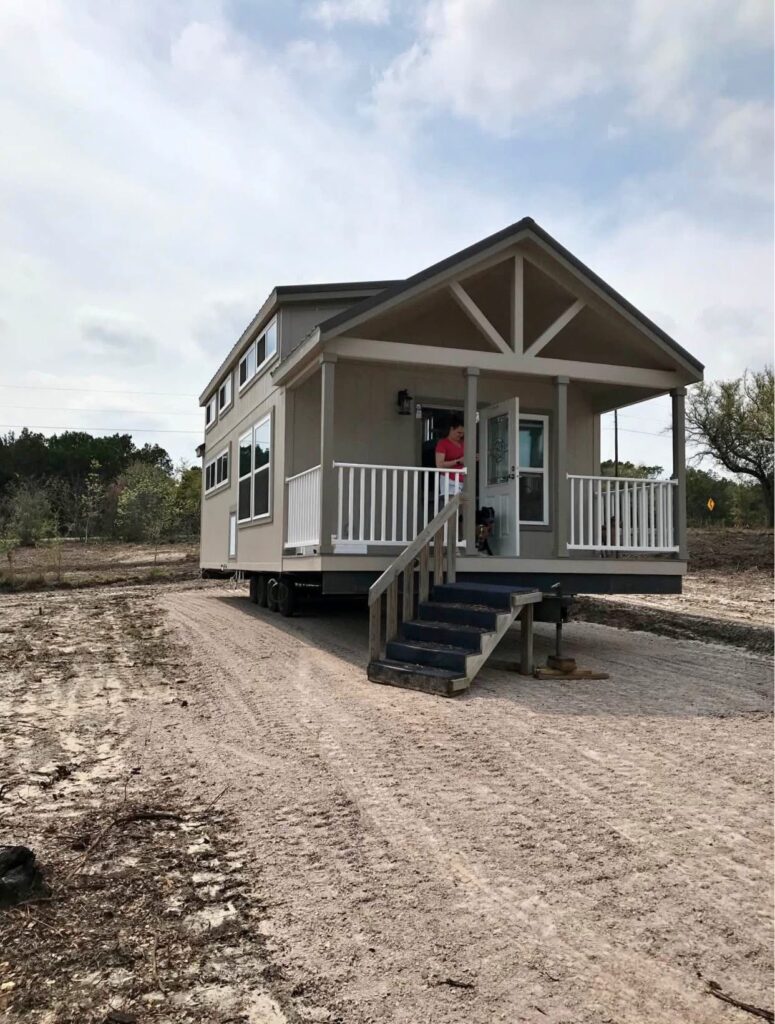 A beige tiny house on wheels with a front porch and stairs sits on a dirt lot. Overcast sky, sparse trees and bushes in the background. A person stands on the porch.