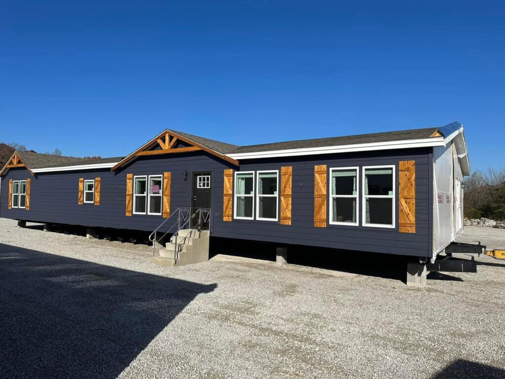 A modern prefabricated home with dark siding and wooden shutters is situated on a gravel lot. It features gabled roofs and large windows, set against a clear blue sky.