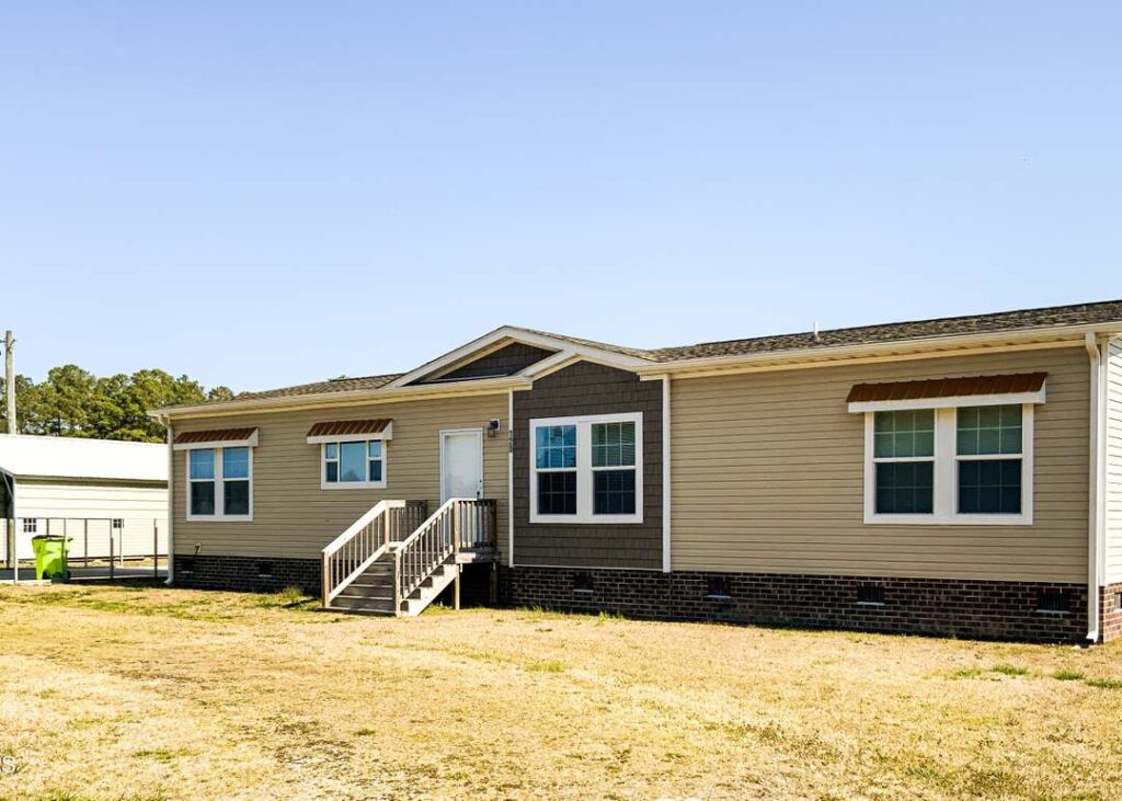 Single-story manufactured home with beige siding and a small porch in a sunny, rural setting. Features multiple windows and a brick foundation.