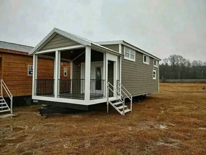 A compact, taupe tiny house with white trim and a covered front porch sits on a field. It has steps leading up and a modern, minimalist design.