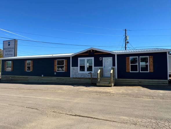 A long, dark blue manufactured home with a metal roof and multiple windows sits under a clear blue sky. A sign reads "Regional Homes."