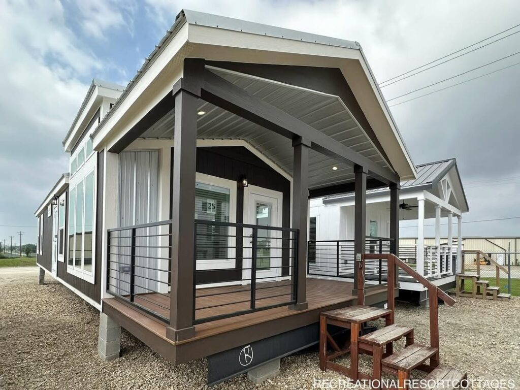 Small modern cabin with a covered porch, wooden steps, and black railings. The exterior is dark with white trim. Cloudy sky in the background.