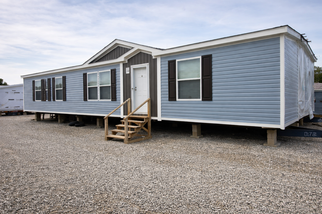 A light blue mobile home with white trim and dark shutters sits elevated on concrete blocks over a gravel lot. Wooden steps lead to the front door.